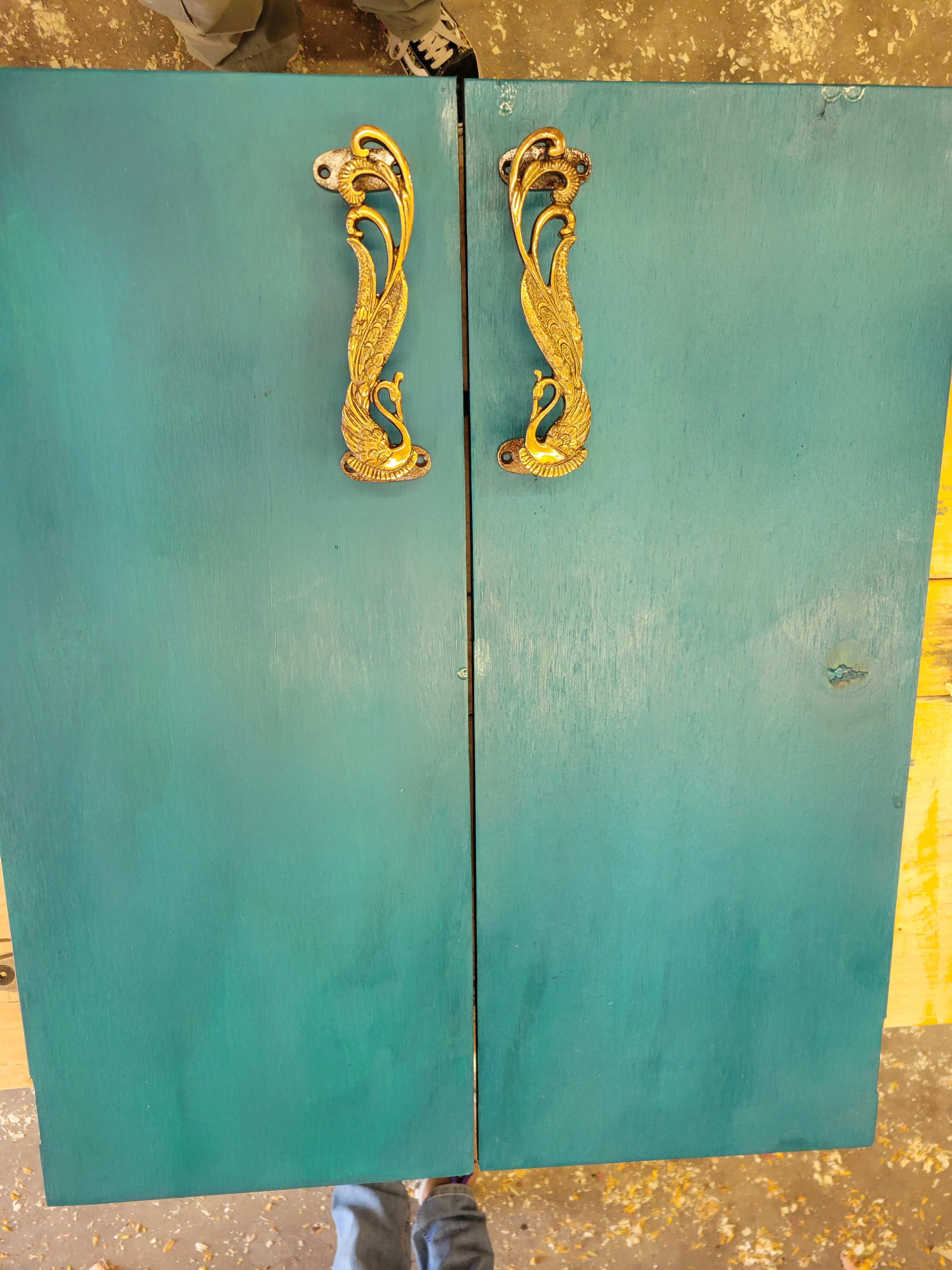 two peacock blue door panels resting on a wooden workbench. there are two brass stylized peacock handles resting near the door edges facing each other.