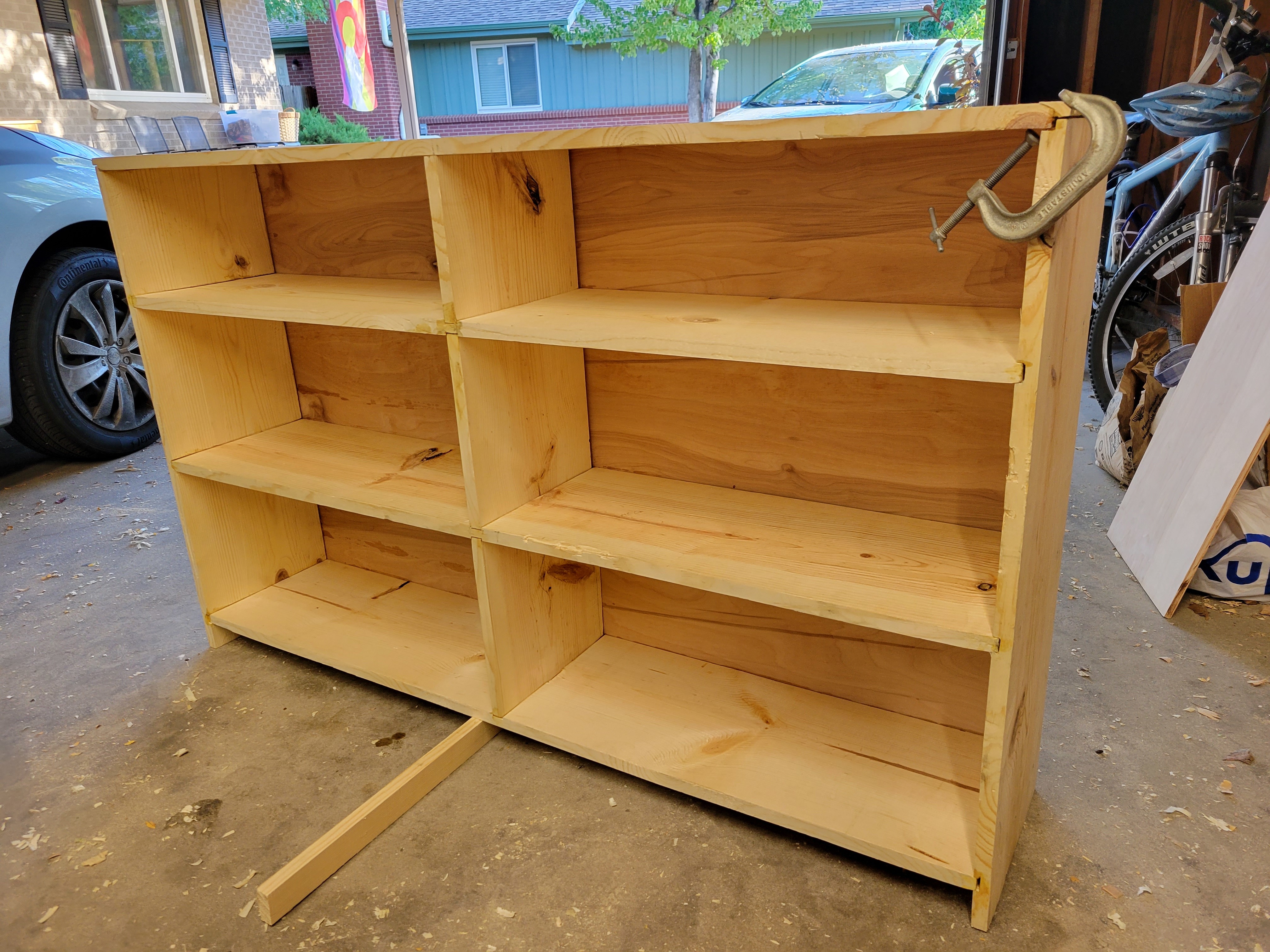 a rectangular, unfinished pine cupboard with two shelves. it looks like a large box and is propped up in the middle on a piece of scrap wood